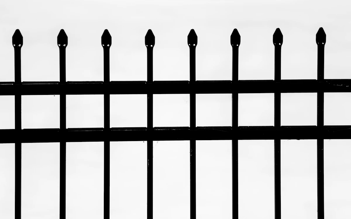 Black metal fence pickets with pointed tops and two horizontal rails against a pale sky